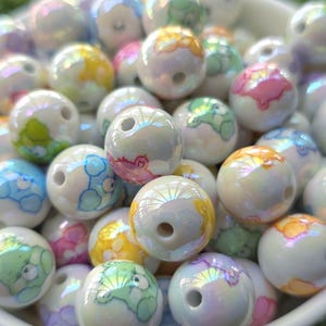 May include: A collection of iridescent white round beads with colorful cartoon bear designs. The beads have a hole through the center and are in a white bowl. The bears are in shades of blue, green, yellow, and pink.