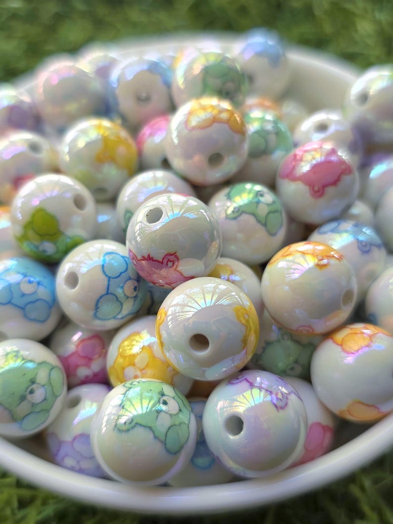 May include: A collection of iridescent white round beads with colorful cartoon bear designs. The beads have a hole through the center and are in a white bowl. The bears are in shades of blue, green, yellow, and pink.