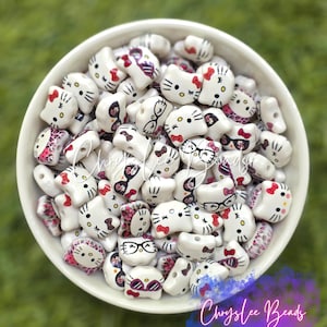 May include: A white ceramic bowl filled with numerous small, white, cat-shaped beads. Each bead features a printed face with black eyes, whiskers, and a yellow nose, with some wearing pink bows or black-rimmed glasses, and others showing pink leopard print.
