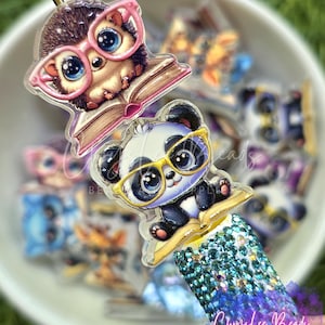 May include: A pen topper featuring two stacked acrylic charms: a brown hedgehog wearing pink glasses reading a book, and a black and white panda wearing yellow glasses reading a book. The base is covered in sparkling light blue rhinestones.