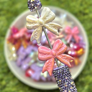 May include: A decorative nail art tool featuring a purple rhinestone-covered handle and a silver rod with a white bow, a pink bow, and a purple rhinestone top. The background is a blurred image of other nail art accessories.