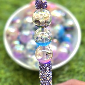 May include: A decorative wand features a sparkling purple handle and a series of clear, iridescent glass orbs. The top orb is a cluster of purple glitter. The orbs have a blue and purple hue.