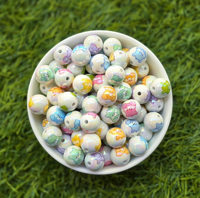 May include: A white bowl filled with iridescent, round beads. Each bead features a colorful cartoon bear design in shades of blue, pink, yellow, green, and orange. The beads are scattered, creating a playful and whimsical aesthetic.