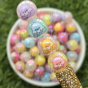 May include: A collection of Care Bear-themed beads in various pastel colors, including pink, blue, and yellow. The beads are round and feature Care Bear characters. The pen is embellished with gold-colored rhinestones.