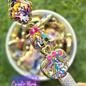 May include: A decorative pen featuring three Sailor Moon character charms with gold-toned metal edges. The charms display colorful anime illustrations and pink bows. The lower section of the pen is covered in small, clear, reflective rhinestones.