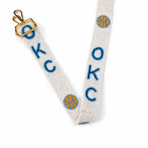 May include: A white beaded strap with the letters "OKC" in blue, and basketball graphics in orange and blue. The strap has a gold-colored clasp. The strap is likely for a bag or purse.