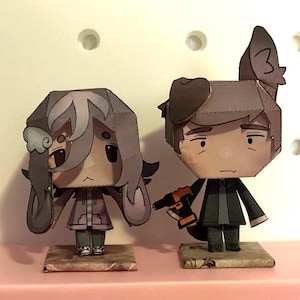 Puede incluir: Dos figuras de papel, una con pelo gris y otra con pelo castaño, están sobre pequeñas bases rectangulares. La figura de la derecha sostiene un taladro. Ambas figuras tienen una expresión triste y están hechas de papel.