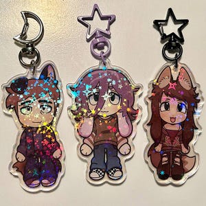 May include: Three clear acrylic keychains featuring cartoon characters with animal ears. The keychains have a holographic glitter effect. Each character is wearing different outfits, including a purple jacket, a brown shirt, and a red jacket. The keychains have star-shaped and crescent moon-shaped clasps.