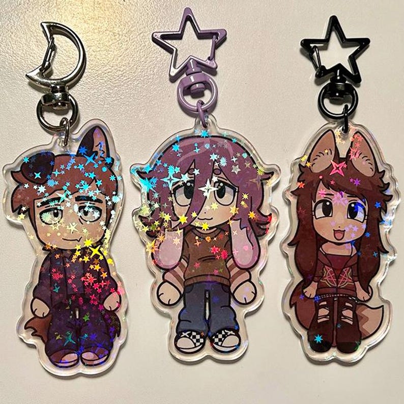 May include: Three clear acrylic keychains featuring cartoon characters with animal ears. The keychains have a holographic glitter effect. Each character is wearing different outfits, including a purple jacket, a brown shirt, and a red jacket. The keychains have star-shaped and crescent moon-shaped clasps.