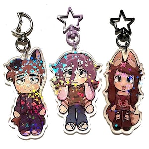 May include: Three cartoon character keychains with glitter accents. Each keychain features a different character with animal ears, wearing various outfits. The keychains have silver, purple, and black star-shaped clasps and a silver crescent moon clasp.