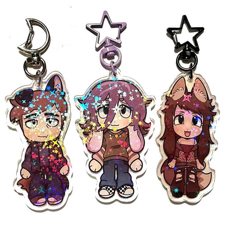 May include: Three cartoon character keychains with glitter accents. Each keychain features a different character with animal ears, wearing various outfits. The keychains have silver, purple, and black star-shaped clasps and a silver crescent moon clasp.