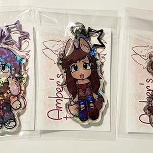 May include: Three clear acrylic keychains featuring anime-style characters. Each keychain has a different character design, with holographic accents. The keychains are packaged in clear plastic bags.