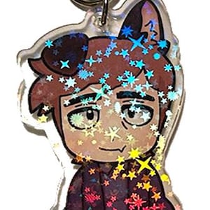 May include: A clear acrylic keychain featuring a cartoon character with brown hair, cat ears, and a purple outfit. The design is decorated with holographic star-shaped accents and a silver metal clasp.