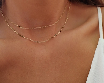 May include: A close-up of a person wearing a gold chain necklace with two strands. The necklace is delicate and features small, round beads.