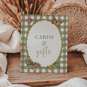 May include: A decorative sign with the words "CARDS & gifts" in elegant script. The sign has a green and white gingham border and features illustrations of butterflies and ladybugs. It is set on a wooden surface.