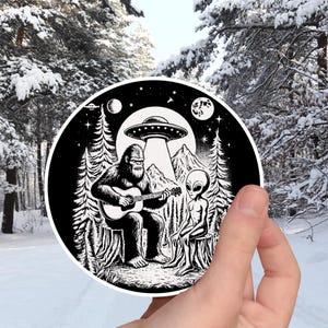 May include: Black and white illustration of a Bigfoot playing a guitar with an alien sitting next to him. The scene is set in a forest with a UFO in the background.