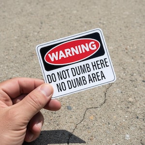 May include: A rectangular sticker with a white background and black border. The top of the sticker has a red oval with the word "WARNING" in white. Below, the text reads "DO NOT DUMB HERE NO DUMB AREA". The sticker is held by a hand.