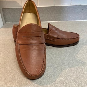 May include: A pair of brown leather penny loafers. The shoes have a classic design with a rounded toe, a strap across the vamp, and a stitched sole. The leather has a smooth texture and a warm, reddish-brown color.