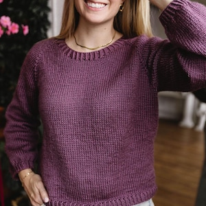 Op de afbeelding: Een pruimkleurige gebreide trui met een ronde hals en lange mouwen. De trui heeft een geribbelde textuur en een getailleerd silhouet. De persoon draagt een gouden ketting en armband. De achtergrond omvat een spiegel en groen.