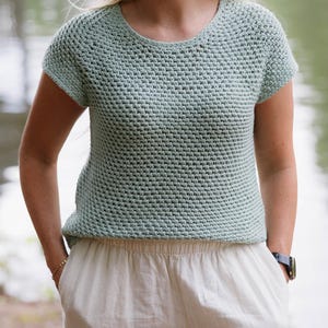 May include: A light sage green short-sleeved top featuring a dense, textured crochet stitch pattern with a rounded neckline. The garment is paired with off-white trousers that have a subtle vertical stripe pattern and an elasticized waistband.