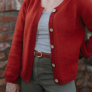 Puede incluir: Un cárdigan de punto color óxido con cuello redondo, mangas largas y cierre frontal con cinco botones circulares de color claro. La prenda se lleva sobre una camiseta de tirantes gris y pantalones verde oliva con un cinturón de cuero marrón.