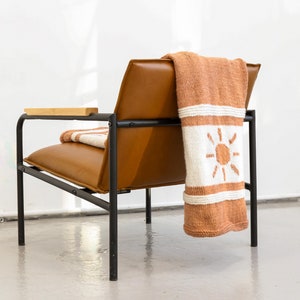 May include: A brown leather armchair with a black metal frame and a wooden armrest. A knitted throw blanket with a white sun design is draped over the back of the chair.