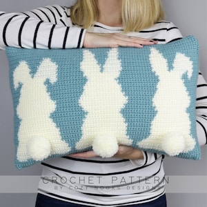 May include: A crochet pillow featuring three white bunny silhouettes on a light blue background. The bunnies have white pom-poms for tails.  "CROCHET PATTERN BY COZY NOOKS DESIGNS" is printed below the pillow.