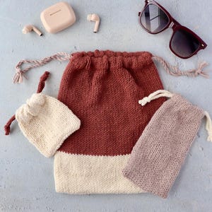 May include: A collection of knitted drawstring bags in various sizes and colors, including a large rust-colored bag with a cream-colored bottom, a small cream-colored bag, and a mauve bag. Sunglasses and wireless earbuds are also present.