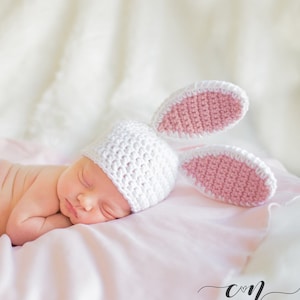 May include: A sleeping baby wearing a white crochet bunny hat with pink inner ears. The baby is lying on a soft pink blanket. The hat has two large, upright ears. The background is a soft, blurred white.