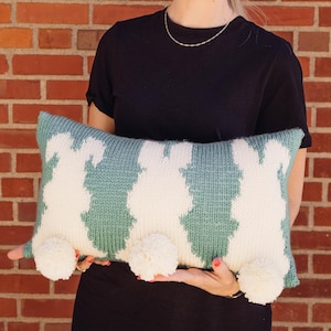 May include: A teal and white knitted pillow with pom-poms on the corners. The pillow has a geometric pattern.