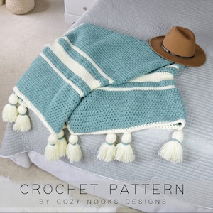 Puede incluir: Una manta de crochet a rayas azul verdoso y blanco con pompones blancos en los bordes. La manta está doblada y drapeada sobre una cama con un sombrero marrón encima. El texto "CROCHET PATTERN BY COZY NOOKS DESIGNS" está en la parte inferior de la imagen.