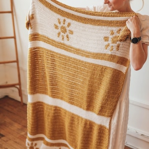 May include: A crocheted throw blanket with a striped pattern in white and mustard yellow. The blanket features a repeating design of stylized suns.