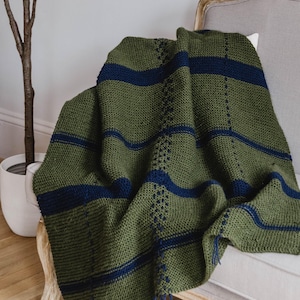 Puede incluir: Una manta de punto verde oliva y azul marino sobre una silla. La manta presenta rayas horizontales azul marino y un patrón decorativo. La manta tiene una franja de hilo azul marino en el borde.
