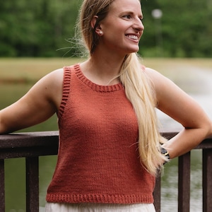 May include: A rust-coloured knitted tank top with a ribbed neckline and armholes. The top is paired with off-white linen shorts. The image is taken outdoors with a blurred green background, suggesting a natural setting.