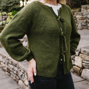 May include: A dark green knitted cardigan with a tie front closure. The cardigan has long sleeves and a relaxed fit.