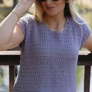 Peut inclure: Un haut en crochet lavande avec un col rond et des manches courtes. Le haut est associé à une ceinture marron et un pantalon blanc. La personne porte des lunettes de soleil et un chapeau de paille.
