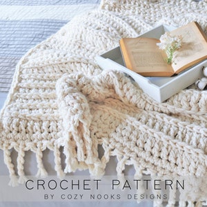 May include: A cream-colored crochet blanket with fringe on the edges is draped over a bed. A book and a tray with cotton balls are on top of the blanket. The text "CROCHET PATTERN BY COZY NOOKS DESIGNS" is at the bottom of the image.