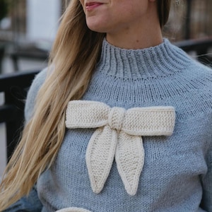 May include: A pale blue knitted sweater featuring a cream-coloured bow. The bow is positioned centrally on the chest, providing a decorative accent. The sweater has a high neckline and a textured knit design.