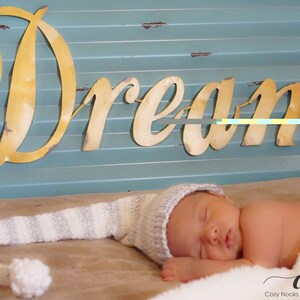 Puede incluir: Un beb&eacute; reci&eacute;n nacido con un gorro de punto blanco y gris a rayas duerme sobre una manta blanca. El beb&eacute; est&aacute; acostado sobre una superficie marr&oacute;n con un letrero de metal dorado que dice "Dream" encima.