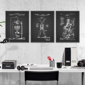 May include: Three black framed prints of vintage patent drawings on a white wall. The drawings are of a glass apparatus, a coffee maker, and a telescope. The prints are hanging above a white desk with a black lamp, a black box, a laptop, a mug, and a camera.