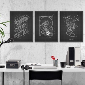 May include: Three black framed prints of computer hardware schematics on a white grid background. The prints are hanging on a concrete wall above a white desk with a black lamp, a black box, a laptop, a mug, a camera, and a black chair.