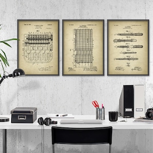 May include: Three framed vintage patent drawings on a white wall above a white desk with a black lamp, a black box, a laptop, a mug, a camera, and a black chair. The drawings are for a typewriter, a grid, and a pen.