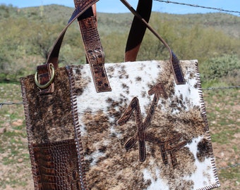 May include: A brown leather tote bag with a white and brown cowhide pattern. The bag has a large, brown leather arrow design on the front. The bag is hanging from a wooden fence post.