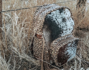May include: A brown and black leopard print backpack with a cowhide flap and fringe detail. The backpack is sitting on a bed of dry grass.