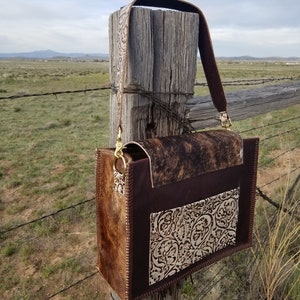 May include: A brown leather handbag with a cowhide flap and a brown leather strap. The bag has a white floral design on the front and is hanging on a wooden fence post.