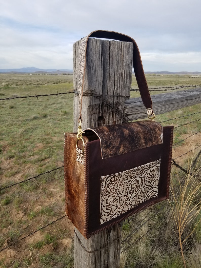 May include: A brown leather handbag with a cowhide flap and a brown leather strap. The bag has a white floral design on the front and is hanging on a wooden fence post.