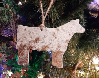 May include: A brown and white cowhide ornament in the shape of a cow. The ornament is hanging on a Christmas tree.