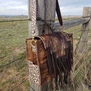 May include: A brown and white cowhide leather handbag with fringe detailing and a white floral patterned strap. The bag is hanging on a wooden fence post.
