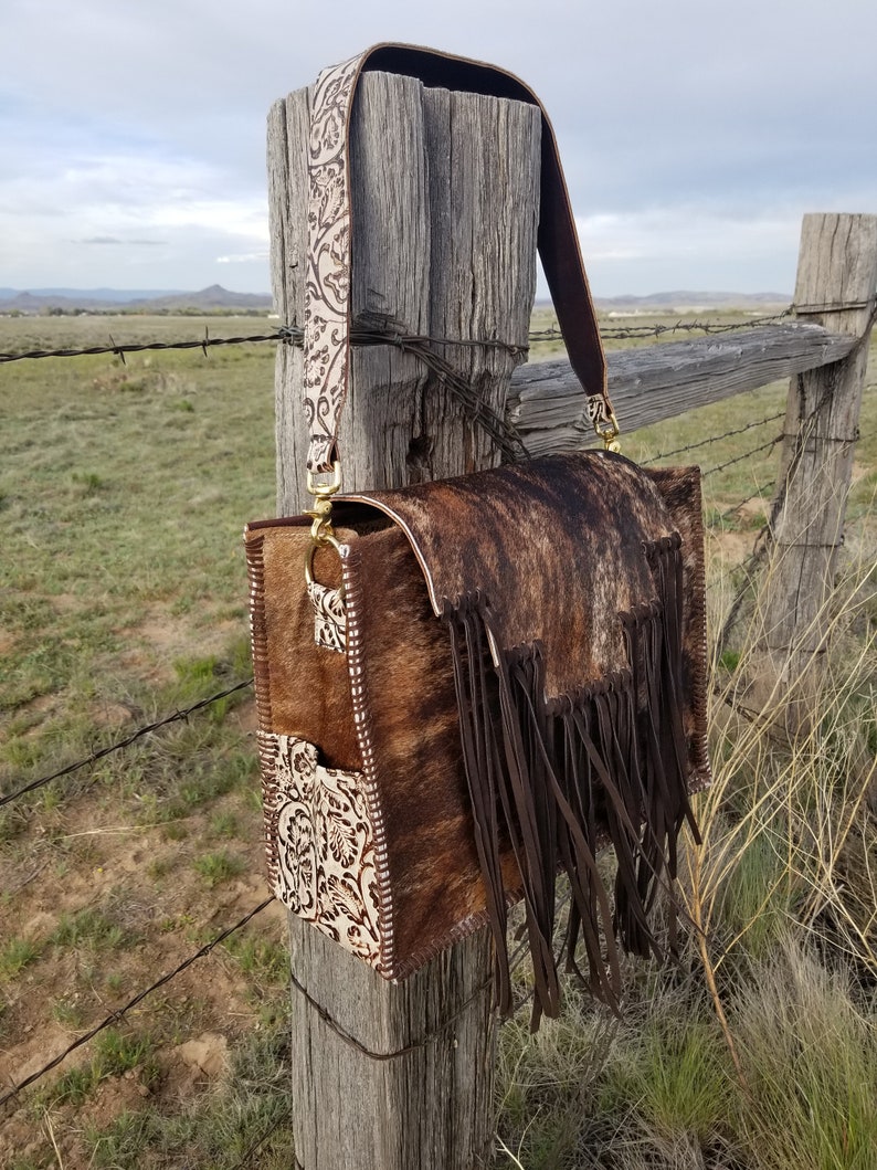 May include: A brown and white cowhide leather handbag with fringe detailing and a white floral patterned strap. The bag is hanging on a wooden fence post.