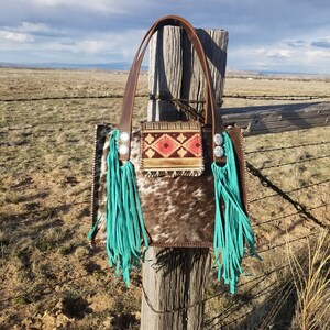 Puede incluir: Un bolso tote de cuero marr&oacute;n con flecos turquesa y un patr&oacute;n geom&eacute;trico colorido en la parte delantera. El bolso est&aacute; colgado en un poste de madera de una valla.